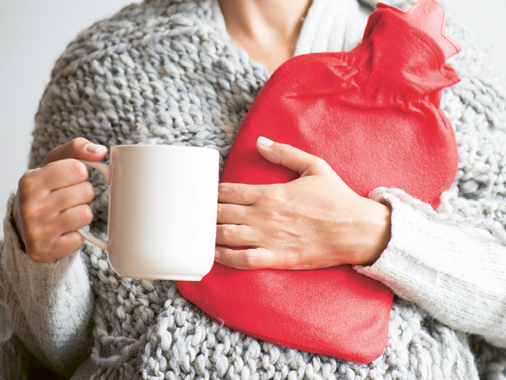 Frau mit grauem Pullover hält eine weisse Tasse und eine rote Wärmeflasche.